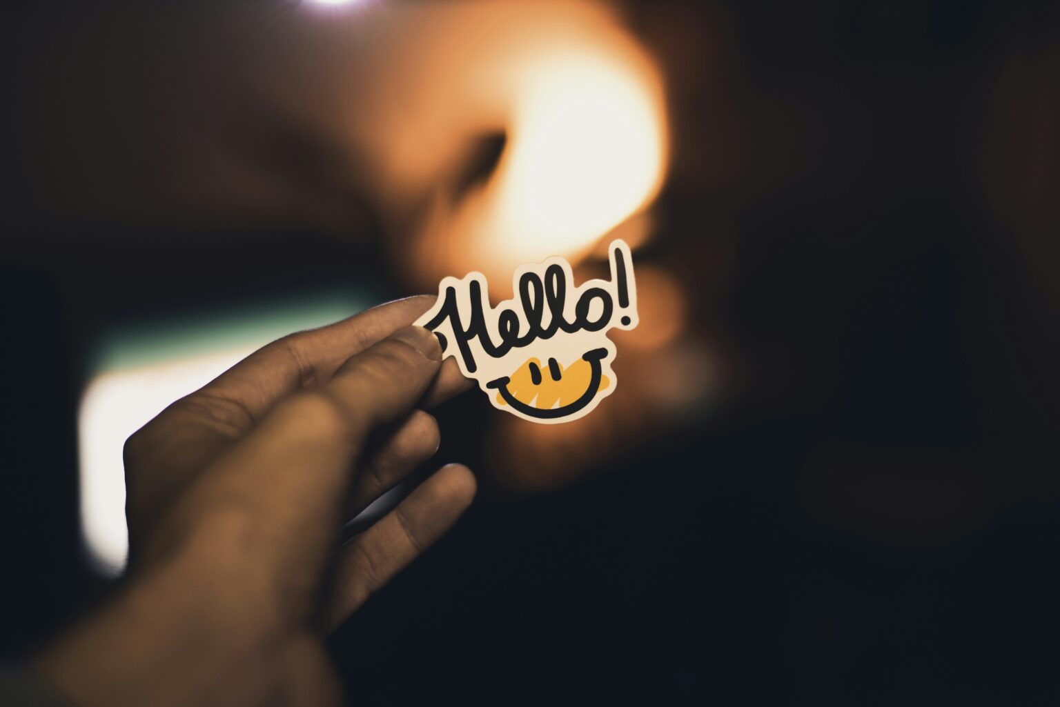 A hand holding up a smiling 'Hello!' sticker against a warm, glowing background